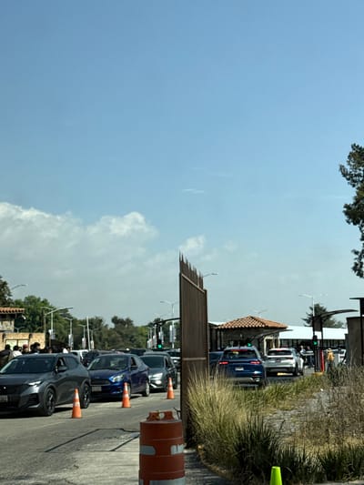 A street with a line of cars is visible in the lower right. Orange traffic cones are placed between the vehicles. A building with a tiled roof can be seen further down the road. To the left of the cars, a fence-like structure is visible, with streetlights lining the street. The sky is predominantly blue with some clouds visible. A tree is partially visible in the upper left corner.