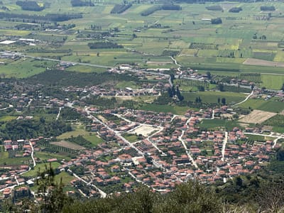 This aerial view shows the town of Ula, Turkey, nestled within a landscape of fields and orchards. The town itself is characterized by closely-packed residential buildings, most with orange-tiled roofs, intersected by a network of streets and roads. A central square, or open area, is visible among the buildings. In the distance, a patchwork of cultivated fields extends into the horizon, varying in color and texture. Roads and smaller paths traverse the landscape, connecting the town to the surrounding agricultural areas. The image is taken from a high vantage point, likely a hillside, as indicated by the presence of vegetation in the foreground. The lighting suggests a clear, sunny day.