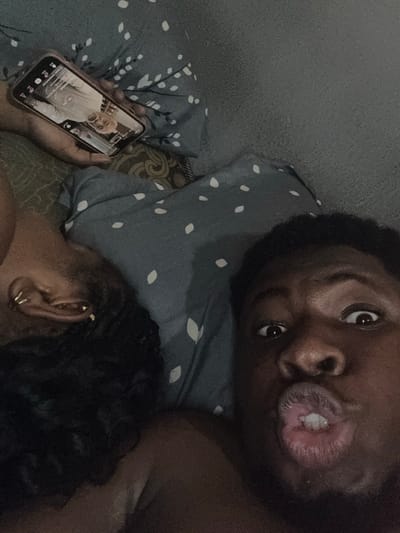 A man and woman lie in bed, the man looking into the camera with pursed lips. The woman’s head is on a pillow with white floral design. A hand holds a cell phone displaying a TikTok video of an older white man. The setting is a room with a gray wall. The location is Gbongan, Nigeria.