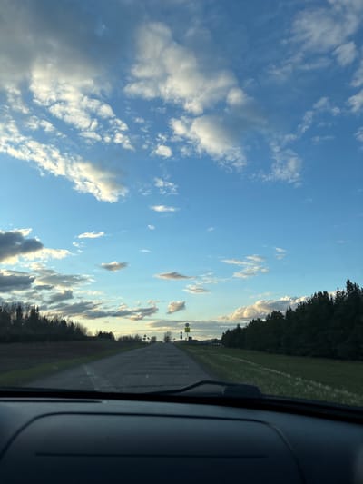 The image captures a landscape view from inside a vehicle, looking towards the horizon. A dark grey section of the car's interior, possibly the dashboard or door frame, occupies the right edge of the frame. A paved road extends from the foreground into the distance, bordered by wide green fields. Dense lines of dark green trees run along the edge of these fields, forming a natural boundary against the bright sky. The sky is a prominent feature, displaying a clear blue expanse interspersed with numerous white and grey cumulus clouds. The quality of light, with the sun appearing lower and casting a gentle glow on some clouds, suggests the time of day is late afternoon or early evening. In the far distance along the road, two yellow diamond-shaped road signs are visible on a pole, though their specific symbols are indistinct due to distance. The scene portrays a peaceful, rural journey through an open landscape. The setting aligns with the provided location context of Aktaziki, Russia. No discernible text is visible within the image.