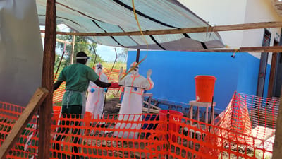 The image captures a scene at what appears to be a decontamination or PPE doffing station in Tchere, Democratic Republic of the Congo, likely operating outdoors under a temporary shelter. Bright daylight suggests it's daytime.

Three individuals are present within a designated area enclosed by orange plastic safety fencing. In the foreground on the left, a person wearing green medical scrubs, a white apron, and a black head covering with safety glasses, stands with their back to the viewer. They are holding a white and black spray device, directing it towards another individual.

This second person, centrally positioned, is fully clad in a white hazmat suit, commonly known as personal protective equipment (PPE), with orange trim. The brand "Tyvek" is visible on their suit, indicating its material. They are also wearing a clear face shield and light blue gloves, and have both hands raised, possibly in a gesture of cooperation for being sprayed or adjusted.

A third individual, similarly dressed in a white hazmat suit with a face shield marked "SAFETY" and blue gloves, is partially visible on the left, slightly behind the first person.

The temporary shelter overhead is constructed from wooden beams and covered with a light-colored tarp, offering shade. In the background, a vibrant blue building wall is visible. To the right, an orange bucket sits on a wooden stool. Text clearly visible on the bucket reads "SALINA," "0.9%," and "DESHABILLAGE," which is French for 'undressing' or 'doffing,' suggesting it's part of a doffing procedure, possibly for a saline solution used in decontamination. The ground beneath the shelter appears to be dirt or mud, with some sparse vegetation in the background. A watermark "Joseph Kisuki" is visible in the bottom right corner.