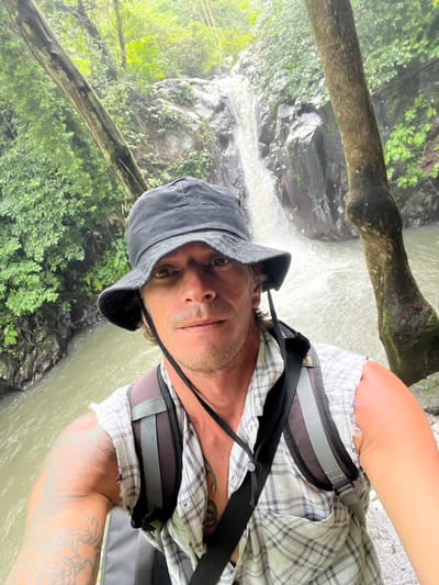 A man in a bucket hat, plaid shirt, and backpack poses, a flowing waterfall serving as the backdrop. Lush green foliage flanks the waterfall on either side, and a fallen tree trunk adds a natural frame to the scene. He looks directly at the camera. The image is rotated counterclockwise.