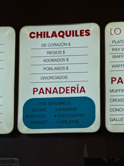 The image shows two menu boards stacked vertically, with a dark background. The left menu lists "CHILAQUILES" with subcategories like "DE CORAZÓN $", "REGIOS $", "ADOBADOS $", "POBLANOS $", and "DIVORCIADOS". The adjacent menu showcases "PANADERÍA" options. It includes "ROL DE CANELA", "SCONE", "BOSTOCK", and "BISQUET", along with "BROWNIE", "CHOCOLATIN", and "OREJITAS" presented as bullet points. Parts of the third menu on top are visible that mention "LO" options like "PLATO", "PAY V", and "WAFFL" and "PA" options like "MUFFIN", "CROISS", "CONCH", and "GALLET".