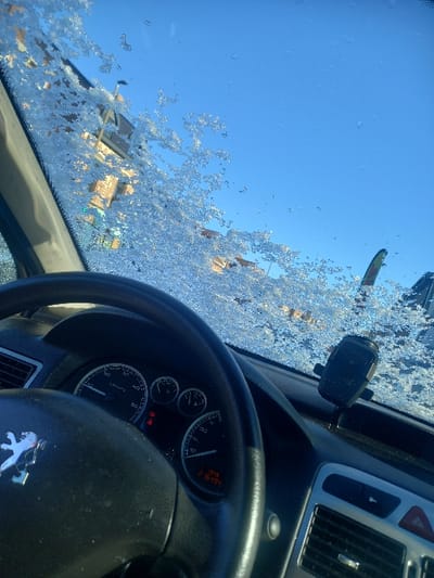 The image depicts the interior perspective from a stationary Peugeot vehicle in Noyelles-sous-Lens, France, looking forward through a windshield partially covered with melting ice and frost. The vehicle's dashboard is visible, featuring a black steering wheel with the Peugeot emblem, and an instrument cluster displaying 0 km/h, the time 13:48, and 216,494 kilometers on the odometer. Multiple red and amber warning lights are illuminated. A black phone mount is attached to the dashboard, and a red hazard warning button is visible.
Through the icy windshield, a clear blue sky is visible above structures. The ice, characterized by crystalline and watery patches, partially obstructs the view of brick or stone multi-story buildings in the mid-ground. A colorful object, possibly a flag or banner featuring yellow and green, is partially visible to the far right. Dried leaves are visible within the ice on the windshield. No persons are present. The scene indicates cold weather conditions with ambient sunlight.