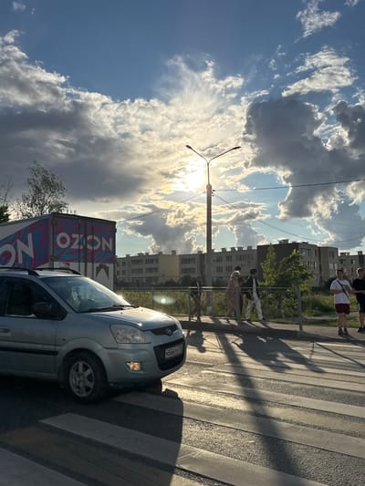 The image depicts a scene on a street in Rastunovo, Russia, under a dramatic sky filled with heavy, textured clouds and bright sunlight peeking through. In the foreground, a silver Hyundai Matrix car is positioned on a crosswalk, its headlights illuminated. The car's license plate is visible, reading "M566KK 750". To the left, a white truck with the "OZON" logo, featuring pink and blue wavy stripes, is parked.
Further back, a sidewalk and a metal railing separate the street from a grassy area. Several pedestrians are walking along the sidewalk. Among them, a group of three individuals, two women and one man, are walking together, with the women in light-colored clothing and the man in darker attire. To the right, two young men are standing, one wearing a white t-shirt and red shorts, and the other in darker clothing. Behind the sidewalk, a row of apartment buildings with multiple floors and windows is visible. A streetlamp with two lights stands prominently near the center of the image. The overall lighting suggests late afternoon or early evening, with long shadows cast across the crosswalk and the ground.