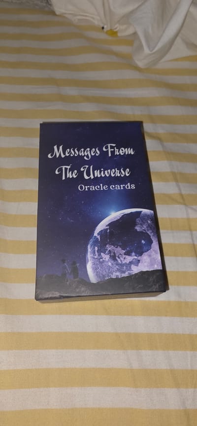 A tarot card box, titled "Messages From The Universe Oracle cards," lies on a bed with yellow and white striped sheets. The box features an illustration of two silhouetted figures standing on a rocky landscape, looking up at a large, detailed planet in a starry sky. The box is angled, showing its spine and the front cover. No information about Lieli, Switzerland, is present in the image.
