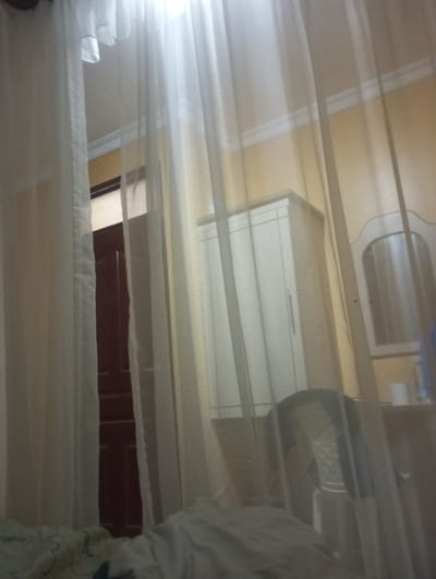 A bedroom view through sheer white curtains reveals a partially visible bed in the foreground. A closed, dark red door peeks through the curtains. An off-white wardrobe and a vanity with a mirror are against a light yellow wall. A chair with a dark jacket draped over it sits near the vanity. The room appears softly lit.