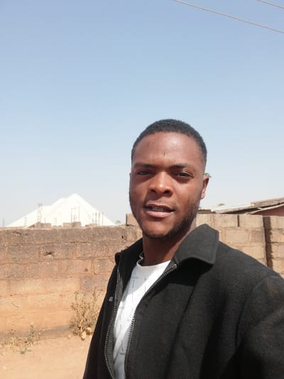 A young adult male of African descent is depicted in a close-up, outdoor portrait taken in Bahit, Nigeria, under a clear, light blue sky. He faces the camera directly, positioned slightly to the right of the frame's center. He has short, dark hair and a sparse beard, and is wearing a black collared jacket with a visible zipper over a white t-shirt. His expression is neutral, with his mouth slightly open.
The background features a weathered, reddish-brown brick wall extending across the midground. Beyond this wall, to the left, a large, light-colored tent-like structure with a pyramidal roof is visible, surrounded by exposed metal rebar, suggesting construction or scaffolding. To the right, behind the wall, a portion of a building with a corrugated metal roof is present. The immediate foreground consists of dry, light brown soil with sparse, withered vegetation. Faint overhead wires are visible against the sky in the upper left quadrant.