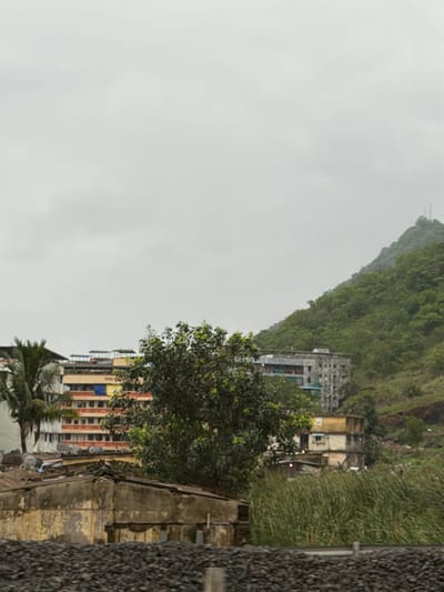 A cityscape with a mountainous backdrop is depicted. In the foreground, a pile of dark, granular material, likely coal or gravel, occupies the bottom of the frame. Above this, a weathered, low-rise building with a textured, yellow facade is visible. Its roof is flat and appears to be constructed from corrugated metal. To the left of this building, a portion of a multi-story apartment building with a white exterior and red balconies is seen, along with a palm tree. Further to the right, a dense, leafy tree stands prominently. In the middle ground, a cluster of multi-story buildings with a colorful facade, featuring red, orange, and yellow sections, dominates the scene. Behind these buildings, a large, green, tree-covered hill rises, with a modern, grey building visible near its base. The sky is overcast with a pale grey hue, suggesting a cloudy day. The overall scene appears to be a residential area within Vasai-Virar, India, characterized by a mix of urban development and natural landscape.