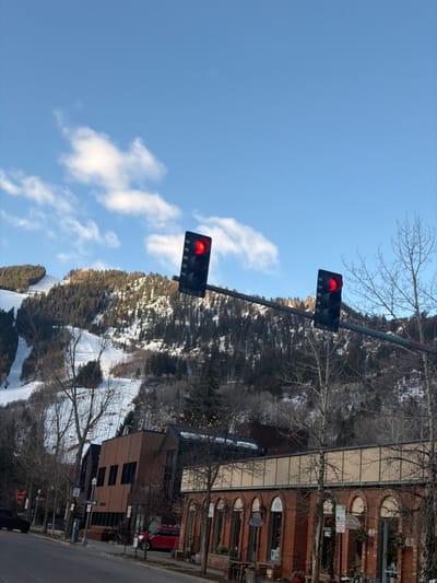 The image displays a street scene in Aspen, Colorado, United States of America, under a clear blue sky with scattered white clouds. Two black, vertically oriented traffic lights are mounted horizontally on an overhead pole, both illuminated red. The street is asphalt and appears dry.

On the left side of the street, a dark-colored vehicle is partially visible, alongside a red "STOP" sign and a "15" MPH speed limit sign. Further along the street on the left, a multi-story building with brown-toned paneling displays "COMPASS" signage. A red car is parked in front of this building. On the right side of the street, a brick building with multiple arched windows and signage, including "TITANIC GAMING," lines the sidewalk. Bare deciduous trees with visible string lights are present along the street and sidewalks.

The background features snow-covered mountains with several visible ski runs cutting through dense evergreen forests. The scene conveys a winter or early spring day in a mountainous resort town. No individuals are visible.