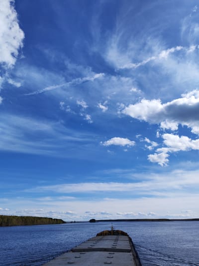 The image depicts a wide-angle view from the stern of a large barge moving across a body of water, likely a river or large lake. The sky dominates the upper portion of the frame, showcasing a vibrant blue hue interspersed with various cloud formations including wispy cirrus and cumulus clouds. A distinct contrail is visible as well. The barge itself is made of concrete. The body of water reflects the sky's color and the surrounding environment. On the left bank of the water body, a dense green forest line runs parallel to the shore. A small island is visible in the distance.
While the image contains elements typical of a waterfront landscape, there is no definitive visual information present in the image to confirm a specific location such as Topornya, Russia.
