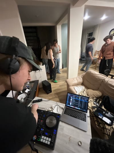 The scene depicts an indoor social gathering, likely in a residential setting in Заозерное Снт, Russia. In the immediate foreground, a fair-skinned individual wearing a dark baseball cap and black over-ear headphones is actively operating a black DJ controller with various illuminated buttons and a circular jog wheel. To their left, a white mug and other small items are visible on a darker surface. To their right, a silver laptop displaying DJ software with a tracklist and waveforms is open on a light-colored wooden table. A beige leather-like sofa with a patterned throw blanket rests behind the laptop, partially obscuring the view.
In the midground, several individuals are standing and interacting within a brightly lit interior space with light-colored walls and wooden flooring. One person in a white tank top and blue jeans holds a glass, conversing with a person in a white top and dark pants. Another person, partially visible, wears a light brown jacket. A black subwoofer is positioned on the floor near a structural column. Further into the room, two more individuals are observed; one in a blue shirt and light jeans walks towards the left, while another in a brown jacket and dark pants stands near a wooden dining table and chairs. A large window with vertical blinds is visible in the background, along with a dark staircase. The overall impression is a casual, active social event.