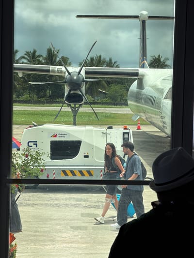 The image shows a view through a window in Del Carmen, Philippines, capturing an airplane on the tarmac and a couple walking towards it. The airplane has a visible propeller and is marked with "Philippine Airlines". The couple is casually dressed. A piece of airport equipment, possibly a generator, is positioned next to the plane, adorned with a "Philippine Airlines" logo and a yellow and black hazard stripe. Palm trees and overcast skies are visible in the background.