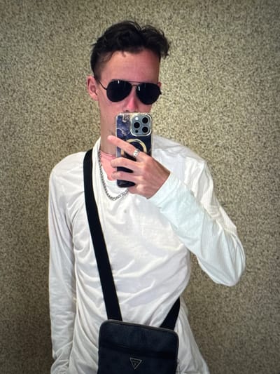 A young man, seen from the chest up, stands in front of a textured, neutral-colored wall. He is wearing a white long-sleeved shirt, aviator sunglasses, and a silver chain necklace. He is holding a smartphone with a marble-patterned case, taking a selfie. A black crossbody bag with a patterned strap is visible across his chest. His left hand, adorned with a ring, is holding the phone. The background appears to be an indoor wall with a speckled texture, possibly in a public space or residence in Porrentruy, Switzerland.