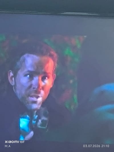 The image shows a close-up, blurry, and somewhat distorted view of a man's face, with significant red and blue lighting on his skin. His hair is dark and appears somewhat disheveled. He is wearing a dark garment. In the lower left corner, the text "xiaomi 13T M.A" is visible, indicating the device used to capture the image and a possible initial. In the lower right corner, the date "03.07.2026 21:10" is displayed. The background is dark and out of focus, with some indistinct colored shapes, possibly from a screen or lighting effect. There is no discernible indication of the location in the city of Орловка, Russia.