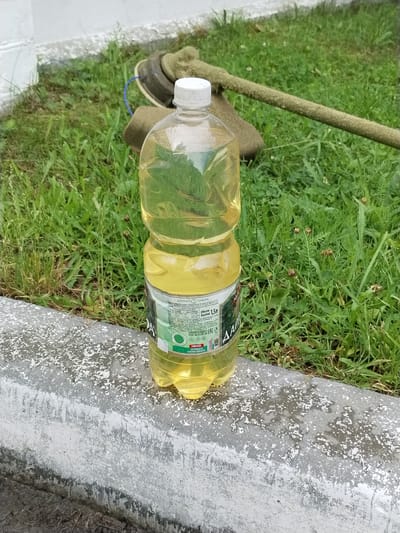 A plastic bottle filled with a light yellow liquid, presumably oil, stands on a concrete curb. The bottle has a white cap and a label with Cyrillic text and a green circular logo. Behind the bottle, a string trimmer is visible, lying in a grassy area with some weeds and dried plants. The background shows more grass and a white structure, possibly a wall or building. The scene appears to be outdoors, possibly in a suburban or rural setting. The bottle's label indicates it contains 1.5 liters and is a product of DARIDA. The text also mentions "НОВИНКА ПОПРОБУЙТЕ В НОВОМ ФОРМАТЕ," which translates to "NEW PRODUCT TRY IT IN A NEW FORMAT." The context suggests that the string trimmer might have been used for maintenance in the area where the bottle is placed. The location, Гожскі сельскі Савет, Belarus, implies a rural or semi-rural administrative area.