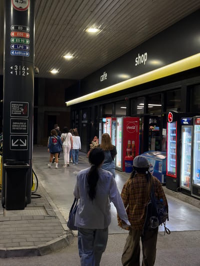 The image depicts a street scene at night, likely a gas station, with a shop and cafe area. The location is identified as Zvornik, Bosnia and Herzegovina.

In the foreground, two individuals are walking away from the camera. The person on the left wears a light-colored long-sleeved shirt and light-colored pants, carrying a dark bag over their shoulder. The person on the right is wearing a plaid shirt, dark pants, and a backpack, with a checkered hat on their head.

A group of four women is walking ahead of them, approaching the shop. They are dressed casually, with one in a blue jacket, another in a white top and light pants, and two more in white tops and jeans.

The environment features a gas pump on the left side of the image, with various fuel types indicated on a vertical panel. The background shows the facade of a building with large windows, displaying rows of illuminated beverage coolers. Signs for "caffe" and "shop" are visible above the entrance. The ground is paved with asphalt and some cobblestone. The lighting is artificial, primarily from the shop's interior, the coolers, and overhead lamps, creating a night-time ambiance.