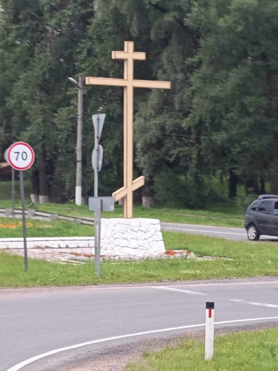 The image depicts an outdoor scene with a large wooden Orthodox cross as the central subject. The cross is light brown and appears to be weathered, standing on a white stone pedestal. To the left of the cross, a circular speed limit sign with "70" in black numerals on a white background within a red border is visible, mounted on a metal pole. Behind the cross and pedestal, dense green trees form the background. A grey vehicle, possibly a hatchback, is parked on the right side of the image, with its rear facing the viewer. The scene is situated near a paved road, with a white line marking the edge. In the foreground, a white post with a red and black marker stands by the roadside. The environment appears to be a rural or suburban area, with green grass and some scattered yellow flowers. The overall impression is of a roadside monument, likely in Russia given the context provided.