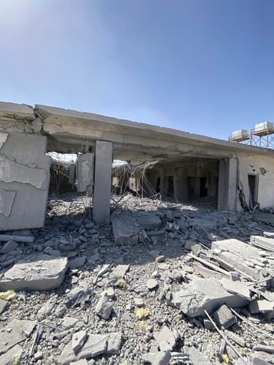 The image displays the extensively damaged concrete structure of a building in Daman, Afghanistan, viewed under a clear, bright blue sky. The upper floor or roof has partially collapsed, with large sections of fractured concrete and exposed, twisted rebar hanging into the interior space. Multiple vertical concrete support columns are visible within the ruin, some still standing, others broken. The ground is covered with a thick layer of debris comprising broken concrete slabs, smaller fragments, dust, and scattered yellowish insulation material. Exposed rebar protrudes from the rubble and damaged structural elements. The interior appears open to the elements, revealing darkened room openings filled with debris. On the far right, atop a raised section of the building or an adjacent structure, two elevated platforms support what appear to be square, white storage tanks. No individuals or active elements are present in the scene.
