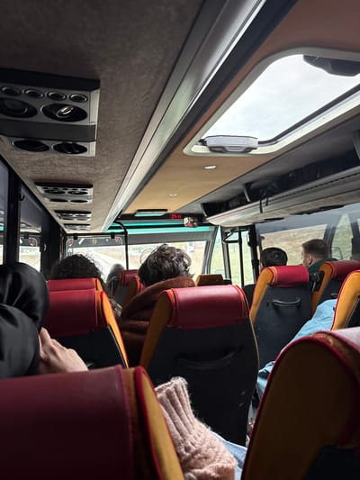 The image captures the interior of a passenger coach in motion within Bańska Niżna, Poland. The view is from a rear-mid passenger seat, looking forward down the aisle. Multiple passengers are seated ahead, observed from behind; their backs are to the camera. The seat directly in front of the camera on the left shows a partial view of a hand near a black garment, while the seat on the right reveals a foot covered in a light pink knitted sock or slipper.

Further ahead, several individuals are visible: a person with dark, curly hair, another wearing a brown, textured jacket, a person with short dark hair, and a person with short light-colored hair wearing a green collared garment. The seats throughout the bus feature red headrests with yellow or orange piping and dark grey fabric bodies.

The ceiling of the coach is lined with grey fabric, incorporating multiple black ventilation and lighting units. An open rectangular skylight with a mesh screen is visible on the right side of the ceiling, allowing ambient light to enter. An overhead luggage rack runs along the right side of the bus, holding dark bags. At the front of the cabin, a digital clock displays "07:14" in red characters. Through the large front windshield and side windows, a glimpse of the exterior environment shows a road, barren trees, and an overcast sky, indicating transit.
