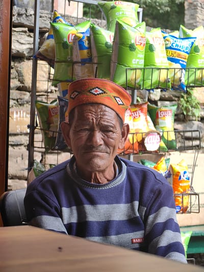 The image shows an elderly man with weathered skin and a mustache, wearing an orange knitted cap with geometric patterns and a dark blue sweater with gray stripes. He is seated at a wooden table, with his upper body visible. Behind him, a rack displays numerous bags of potato chips, primarily Lay's brand, with some bags featuring the "Aloo Bhujia" and "Masala" labels. The background includes a stone wall, indicating an outdoor setting. The overall impression is of a vendor or shopkeeper in Kothi, India, with his merchandise prominently displayed.