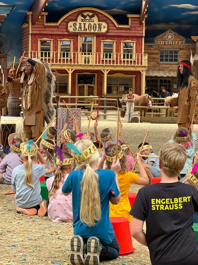 This image captures an outdoor event or performance set against a Wild West-themed backdrop, likely in Collendoorn, Netherlands. In the foreground, a group of children are seated on the ground, facing away from the camera, dressed in costumes and wearing feather headbands, some holding sticks. A boy in a black t-shirt with yellow lettering that reads "ENGELBERT STRAUSS" is prominently visible on the right. In the mid-ground, two individuals dressed as Native Americans are standing, holding sticks. Behind them, a western-style building with a "BOOTHILL SALOON" sign and a "SHERIFF" office is visible. A woman in a cowboy hat stands near a horse. The ground is covered with sand and confetti.
