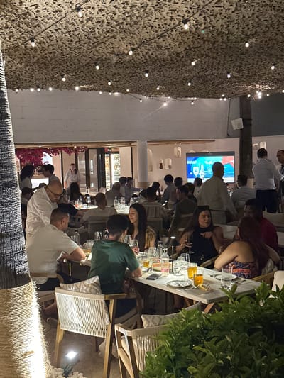 The image depicts a lively outdoor dining scene at a restaurant in Playa Del Sol-Villacana, Spain. The setting is illuminated by string lights draped across a camouflaged overhead structure, and the ambiance suggests an evening gathering. A prominent tree trunk wrapped in rope is visible on the left, adding a natural element to the scene.

Numerous patrons are seated at tables, engaged in conversation and enjoying meals. Many tables are set with plates, glasses, and bottles, indicating active dining. In the background, a large screen displays imagery, possibly a sporting event or advertisement. Restaurant staff can be seen serving guests. The overall environment is that of a bustling restaurant, with people enjoying their time in a social and convivial atmosphere.