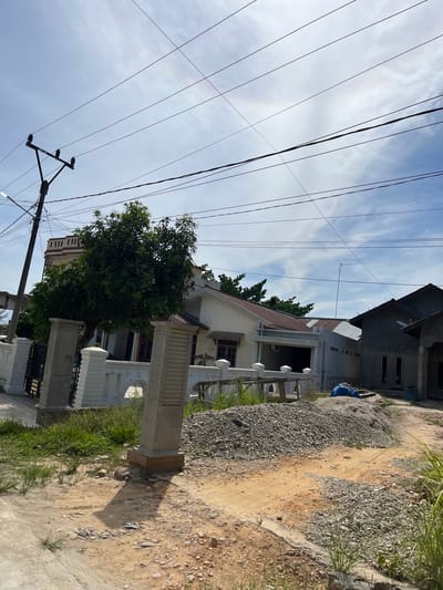 The image depicts a residential scene in Dalam, Indonesia, under a partly cloudy sky. The perspective is from a low angle, looking up towards a jumble of overhead power lines that crisscross the frame. In the foreground, a dirt path with scattered gravel leads past a white fence with decorative pillars and a gate. To the left of the path is a large, leafy green tree, casting a shadow on the ground.

Behind the tree and fence, a cream-colored house with a brown-tiled roof is visible. Its facade features several windows and a covered porch area. To the right of this house, another building is under construction, with its concrete walls and unfinished roof evident. A blue tarp is draped over some materials near the construction site.

The overall impression is one of a developing neighborhood, with a mix of completed homes and ongoing construction. The time of day appears to be late morning or early afternoon, given the bright sunlight and the shadows cast. The weather seems fair, with the scattered clouds suggesting a pleasant day. There is no visible text in the image.