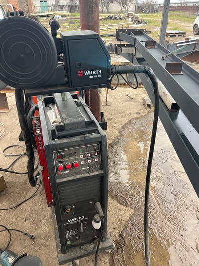 The image depicts an outdoor industrial or fabrication scene in Zlatovrah, Bulgaria. The primary subject is a two-part WÜRTH MIG 500 SYN welding system. The upper unit is black, featuring a visible black welding wire spool on its left side. The lower unit, also black and on wheels, displays a control panel with red knobs and digital readouts of "280" and "283," and is labeled "WR-82 HIGH-FREQUENCY WATER TANK." A thick black corrugated conduit connects the two WÜRTH units and extends to the right. The ground is uneven, composed of dirt and mud, featuring numerous puddles. To the right, a large, dark-colored, fabricated metal beam structure extends diagonally, supporting smaller metal sections and a light-colored square tube. In the lower-left foreground, a portion of a green angle grinder is visible on the ground. The background reveals a workspace with various metal components, rusted vertical poles, scattered debris, and bare trees. A brick building with a light-colored door and a partial white van are visible in the far background. No individuals are present in the scene.