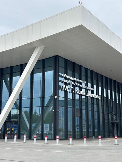 The image shows the exterior of a modern airport terminal building with large glass windows and a white, angular roof structure. White text on the building reads "Международный аэропорт Ижевск имени М.Т. Калашникова" which translates to "Izhevsk International Airport named after M.T. Kalashnikov." The airport is located in the village of Starye Martyanovo, Russia. In front of the building, there are several bollards with red and white stripes. The sky is overcast.