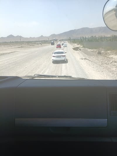 The image shows a convoy of vehicles traveling on a dusty, unpaved road in a mountainous region, likely in Kyrgyzstan given the provided location context of Жаны-Кыштак. The perspective is from inside one of the vehicles, looking forward through the windshield. The road is wide and appears to be a dirt track with loose gravel. On either side of the road, there is sparse vegetation and the foothills of mountains are visible. The sky is bright and appears to be clear, suggesting it is daytime.
Several vehicles are visible on the road, including white sedans, a red car, and possibly a truck further down the line. One white car in the foreground has a license plate with Cyrillic characters. The convoy suggests movement and a journey. The overall impression is one of travel through a remote and somewhat arid landscape. The interior of the vehicle shows a dashboard on the right side of the frame, with a grey textured surface. The reflection of the sky and the road can be seen on the windshield. There are no people clearly visible within the vehicles. The activity is simply travel along a road in a rural, mountainous environment.