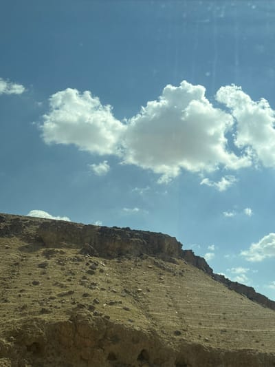 The image captures a low-angle shot of a desert landscape near Arad, Israel. Dominating the right side is a steep, weathered rock formation, its surface exhibiting a rough texture with scattered small rocks and vertical striations. The color is a muted brown, indicative of the arid environment. To the left, the sky is a light blue, punctuated by fluffy white clouds. The composition suggests a contrast between the solid, earth-bound rock and the ephemeral, airy clouds. No people or signs of civilization are directly visible, emphasizing the natural, desolate beauty of the desert landscape.