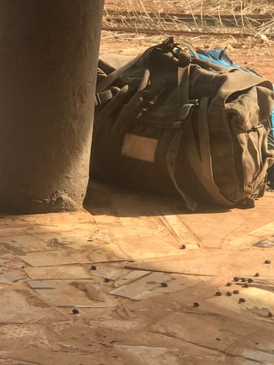 In Okura, Nigeria, the scene depicts a segment of an indoor or semi-sheltered area transitioning to an outdoor environment. A large, olive-drab canvas duffel bag with straps and a rectangular identification patch rests on a floor composed of irregularly shaped, light brown, flat pieces, possibly cardboard or thin wood, laid over a dusty surface. To the left of the bag, a thick, cylindrical column of a similar light brown, dusty material, likely concrete or earth, rises vertically. Its base is rough and crusted with accumulated dust and debris. Scattered across the light brown floor are numerous small, dark, oblong pellets, consistent with animal droppings. In the background, beyond the duffel bag, blurry dry, light-colored vegetation and reddish-brown soil are visible under bright daylight. No individuals, actions, or direct interactions are observable.
