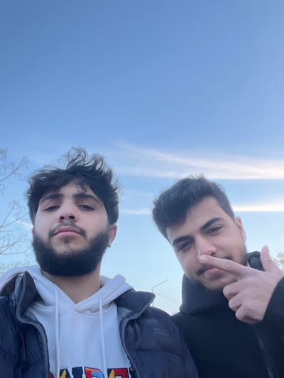 The image displays two young adult males captured in a close-up, selfie-style photograph outdoors, set against a clear blue sky with faint wispy clouds. The location is the city of Qudsaiya, Syria.

The male positioned on the left has dark, tousled hair, a full dark beard, and a neutral facial expression, looking directly forward. He is dressed in a dark, possibly navy blue, puffy winter jacket worn over a white hooded sweatshirt displaying colorful lettering on the front.

The male on the right has dark, neatly styled hair and light facial hair. His left index finger is placed against his nose, and his left thumb is extended upwards, while he looks towards the camera with a slight smirk. He wears a dark, possibly black, hooded garment.

In the background, bare tree branches are visible on the far left of the frame. A light-colored utility pole or street light support structure is faintly discernible in the distant mid-ground, slightly to the right of the center.