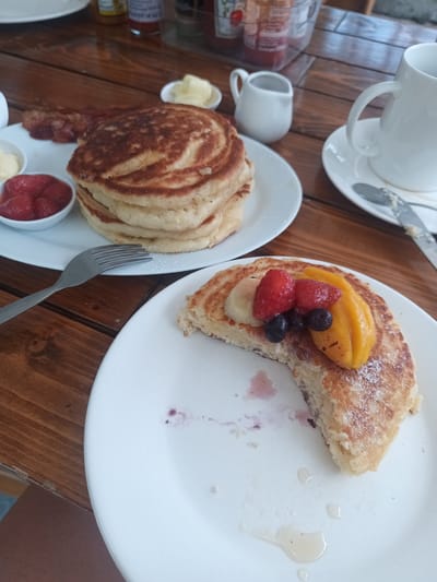 The image depicts a breakfast scene likely taken in Hua Hin City Municipality, Thailand. The setting is a wooden table, with two white plates. The first plate holds a stack of pancakes served with a side of strawberries and butter, accompanied by a fork. The second plate contains a partially eaten pancake topped with banana slices, strawberries, blueberries, and mango. A drizzle of syrup is also visible on the plate. In the background are a white mug with a saucer, a creamer, and various condiment bottles. The composition suggests a breakfast setting in a restaurant or cafe.