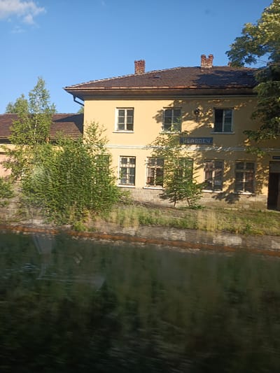 The image shows a two-story building with a yellow facade, likely a train station, in Chelopech, Bulgaria. The building has a red-tiled roof and several windows. A blue sign with Cyrillic text is visible on the facade. Trees and foliage surround the building, and a railway track runs alongside it. The scene appears to be set during the daytime, under a clear blue sky. The image is slightly blurred, possibly due to motion, suggesting it might have been taken from a moving vehicle. The overall impression is one of a quiet, possibly rural, setting. The text visible on the sign appears to be "ЧЕЛОПЕЧ", which is the name of the location.