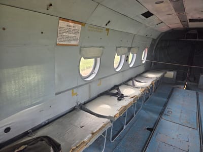 The image shows the interior of an old, possibly abandoned, aircraft cabin. The cabin features rows of basic, bench-like seats with worn upholstery and visible seatbelts. The walls are painted a light gray, and there are several circular windows with white fabric covers. Yellow rectangular markings are visible on the walls, and one sign in Russian with English text "CUT-OUT IN THIS AREA" is present. The floor is a metallic blue with a textured pattern. Some of the seats appear damaged, with exposed foam padding. In the background, the cabin opens into a darker, unfinished section of the aircraft. The overall impression is one of disuse and deterioration. The location is identified as Bukharovo, Russia.