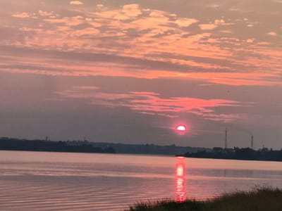 A serene sunset is captured over a tranquil body of water, likely a river or lake. The sky is painted in hues of pink and orange, with streaks of clouds stretching across the horizon. The sun, a vibrant red orb, is partially visible, casting a shimmering reflection on the water's surface. The distant shoreline features a silhouette of trees and buildings, with a few industrial structures and communication towers visible. The water itself is calm, with gentle ripples creating subtle patterns. The foreground shows a grassy bank with tall reeds. The overall atmosphere is peaceful and tranquil, evoking a sense of calm.