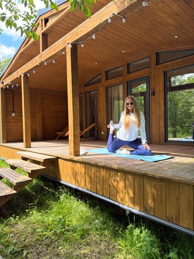 The image, set in Vozhoiskii, Russia, captures a woman on a wooden deck outside a modern cabin. The woman, with long blonde hair, wears a white long-sleeved top, blue leggings, and sunglasses. She is positioned on a light blue yoga mat, performing a yoga or stretching pose that resembles a low lunge or pigeon variation.

The cabin features a distinctive design with multiple large, dark-framed windows reflecting green trees and a blue sky. Its exterior is clad in warm-toned wooden planks. The expansive wooden deck where the woman is located is also made of similar timber. Beneath the elevated portion of the deck, string lights with globe bulbs are strung along wooden support beams, and further wooden structures are visible.

The scene is bathed in bright natural sunlight, indicated by clear shadows, suggesting a sunny day, likely mid-morning to afternoon. Lush green grass and foliage surround the deck, highlighting the natural environment. The number "17" is visible on the cabin wall next to a door on the deck. The overall impression is one of tranquility and connection with nature, possibly at a retreat or vacation home.