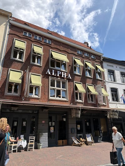 The image depicts the building facade of "ALPHA" restaurant, located in Roermond, Netherlands. The building is constructed with red brick and features multiple windows with light green awnings. The name "ALPHA" is prominently displayed in white lettering on the facade. In front of the building, there is an outdoor seating area with tables and chairs. A woman is walking on the sidewalk in the foreground, carrying a black shopping bag. The street is paved with bricks, and there is a light pole visible. The sky is blue with some clouds. Adjacent to the "ALPHA" building is a white building with arched windows, indicating it is part of the same streetscape. A Dutch flag is visible on the white building. The scene suggests a commercial district with a restaurant open for business.