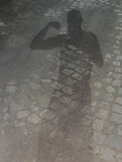 The image is a shadow of a person on a cobblestone street, which appears to be located in the city of Gorna Malina, Bulgaria. The shadow is cast on the ground, which is made up of small, irregular stones arranged in a pattern. The lighting suggests it is either early morning or late evening. The shadow shows a person standing with their left arm bent, as if flexing a bicep. The shadow is dark and indistinct, making it difficult to discern any specific details about the person or their clothing. The surrounding environment is not clearly visible, but the cobblestone pattern suggests an outdoor urban setting.