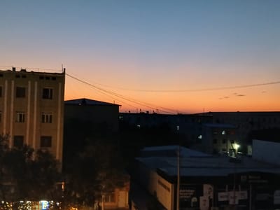 The image depicts a twilight scene over the city of Qorowul, Uzbekistan. The sky transitions from a pale blue at the top to a vibrant orange and pink gradient near the horizon, indicating the sunset or sunrise. Silhouetted against the colorful sky are several buildings, including a prominent multi-story structure on the left with visible windows and vertical decorative elements. In the background, more buildings and structures are discernible, some with illuminated windows. Electrical wires are strung across the sky, a common urban feature. The foreground shows darker silhouettes of trees and some illuminated signs, suggesting commercial activity. The overall atmosphere is calm and quiet, characteristic of a city at dusk or dawn. There are no people or animals clearly visible in the image.
