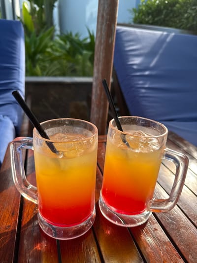 Two frosted glass mugs filled with a layered red and orange beverage sit on a wooden table. Each mug contains ice cubes and a black straw. In the background, out-of-focus blue lounge chairs and lush green foliage are visible. The scene appears to be outdoors, likely in a resort or hotel setting in Cam Lâm, Vietnam, suggested by the tropical vegetation and leisure furniture. The lighting indicates it is daytime, with shadows cast on the table.