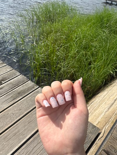 The image shows a person's hand with light pink nail polish featuring small blue and pink flecks. The hand is positioned over a wooden deck. To the left of the hand, there is a body of water with tall green grass growing at its edge. The sun appears to be shining, casting shadows on the deck and creating highlights on the water. The scene suggests an outdoor setting, possibly a park or a waterfront area. The wooden planks of the deck are weathered and laid in parallel lines. The water is calm, with ripples visible on its surface. The grass is lush and appears to be swaying slightly. The lighting and shadows indicate it is likely daytime, possibly late afternoon given the angle of the sun. The overall impression is one of a serene and natural environment. No text is visible in the image.