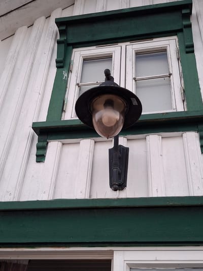 This is a close-up, low-angle shot of the exterior of a white wooden building with decorative dark green trim. A black outdoor lantern with a glass globe is mounted to the wall to the left of a double-paned window. The window is framed by the dark green trim, which also features decorative wooden brackets. The building's siding is white clapboard, with some visible weathering and wear on the paint. The overall impression is of an older, possibly historic, building in a coastal or nautical setting. This image was taken in Åsgårdstrand, Norway.