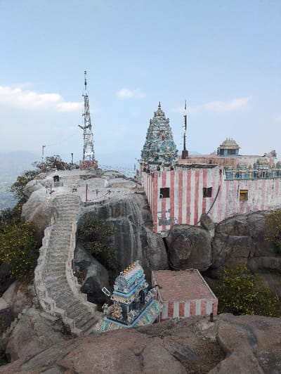 This image displays a temple complex situated on a rocky hilltop in Kadaladi, India. The scene is dominated by grey rock formations under a light blue sky with scattered white clouds. A prominent multi-tiered Hindu temple building, painted with vertical red and white stripes, stands on the higher ground. This building features multiple rectangular windows, one with a yellow frame, and is topped by an ornate gopuram-style tower with intricate blue, white, and gold carvings. To the left and slightly behind this main structure, a tall, lattice communication tower with multiple antennae is erected on another rocky peak. A winding stone staircase with patterned balustrades ascends the rocky slope from the lower left, leading towards the main temple area. Below the main temple, a smaller, similarly ornate shrine with blue, white, and gold carvings is positioned next to a rectangular building with vertical red and white stripes and a reddish-brown tiled roof. Sparse green foliage and bushes with yellow flowers grow among the rocks. No individuals are present. Distant hills are visible in the background under a hazy sky.