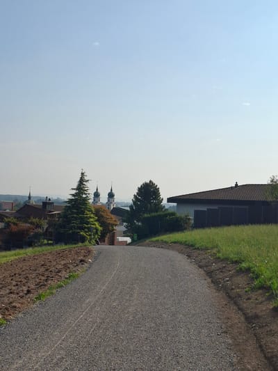 The image captures a view looking up a gravel driveway bordered by a freshly tilled field on the left and a grassy embankment on the right. In the background, a modern building with a dark-tiled roof and gray walls is visible. Beyond that, a cluster of buildings, including a church with two distinct onion domes and spires, is situated on a slight rise. The sky is a clear, pale blue with a few wispy clouds. The overall scene suggests a rural or semi-rural setting, likely in the vicinity of Eschenbach (LU), Switzerland, given the architectural style of the church.