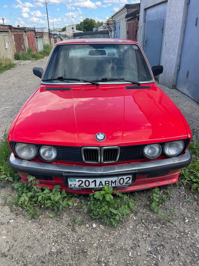A red BMW is parked in front of garages in Michurinsky, Russia. The car is overgrown with grass and weeds. The license plate reads "201 ABM 02". The sky is cloudy.
