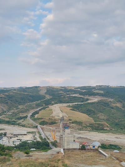 This is an elevated, eye-level, full shot of a construction site in Muğanlı, Azerbaijan, against a backdrop of rolling, tree-covered hills and a partly cloudy sky. The foreground consists of dry, yellowing grass and a dirt path. In the mid-ground, a concrete structure, likely part of a bridge or dam, is under construction, with scaffolding and cranes visible. To the left of the structure, a road winds through the landscape, with a few vehicles on it. Further left, an industrial area with buildings and parked machinery is present. The background features the undulating, green-covered hills of Muğanlı, with some roads and structures visible on their slopes. The sky is a mix of pale blue and white clouds.