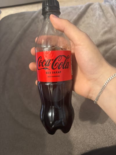 A close-up image displays a light-skinned right hand holding a small, clear plastic Coca-Cola bottle. The hand's fingers are wrapped around the bottle, with a silver-colored curb chain bracelet visible on the wrist. The bottle features a black screw-top cap and a red label with "Coca-Cola" in white script. Below the brand name, Cyrillic text reads "БЕЗ ЗАХАР" (without sugar) and "БЕЗ КАЛОРИИ" (without calories), indicating a zero-sugar variant. The bottle contains a dark brown liquid consistent with Coca-Cola. The background consists of a soft, textured, greyish fabric material, with no distinguishing features. The scene is depicted in Borisovo, Bulgaria, as specified.
